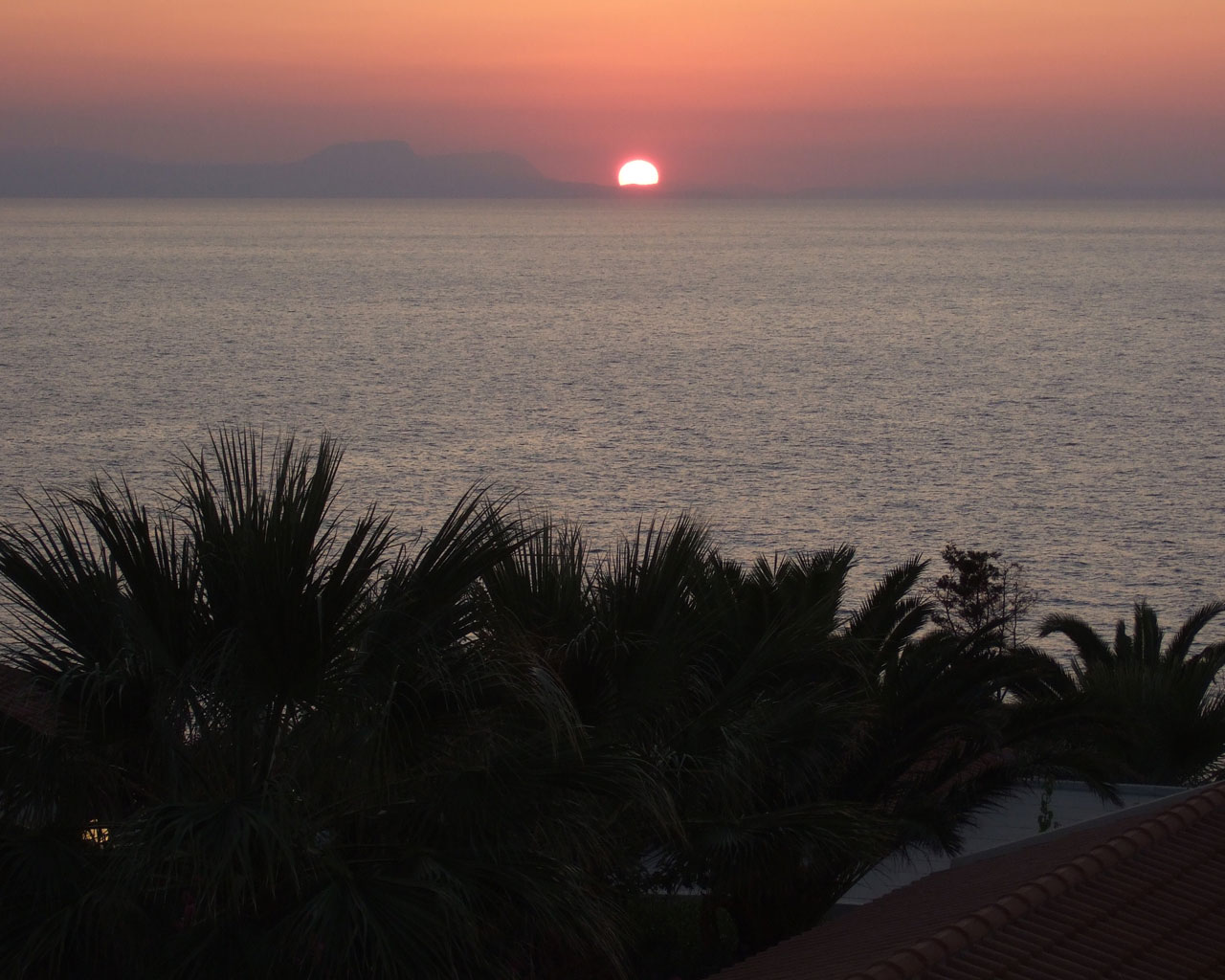 Крит. Вечерний вид с балкона. Солнце
      опускается за гору в море.